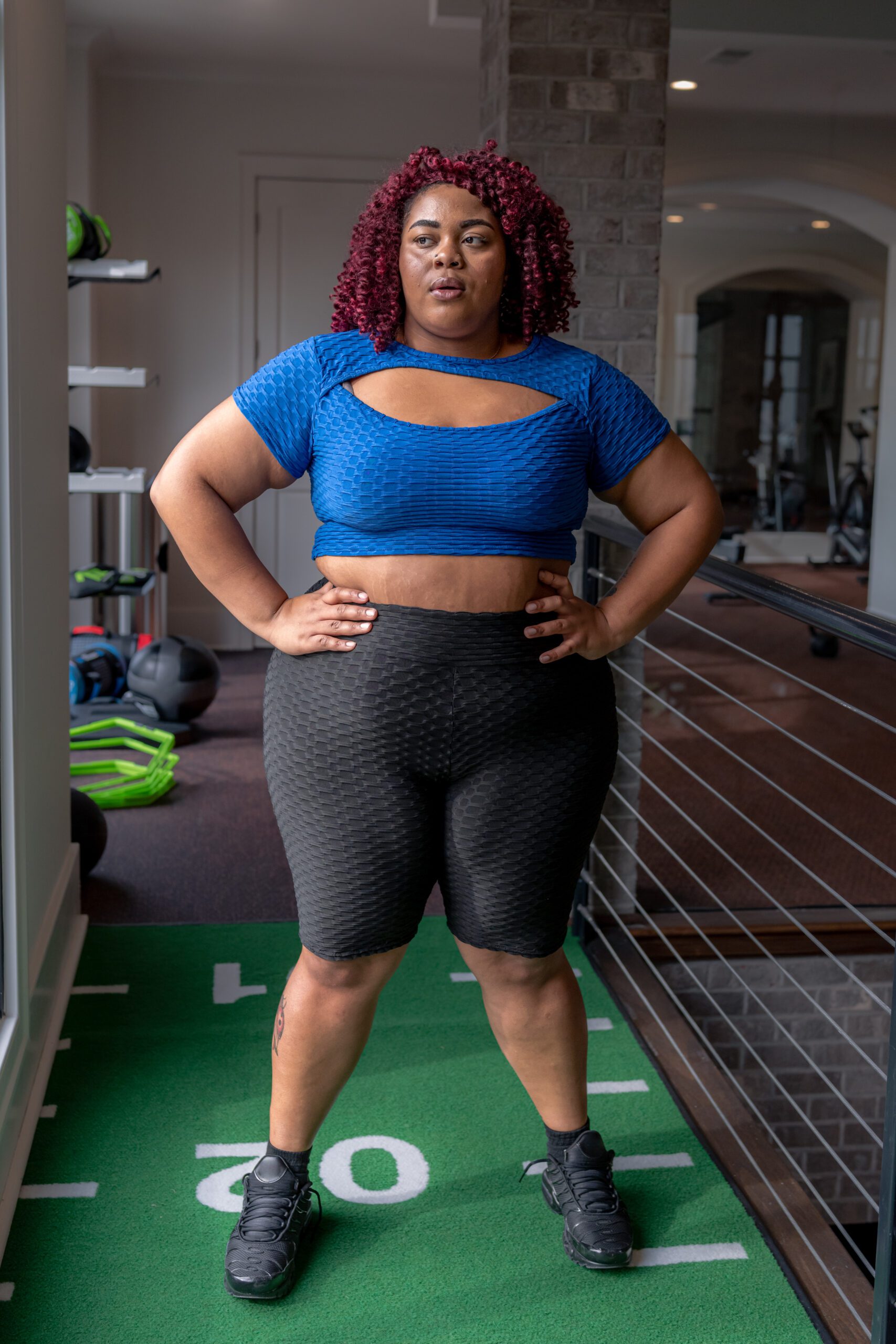 A multiracial woman stands in a gym with her hands on her hips and takes a break from exercising to catch her breath.